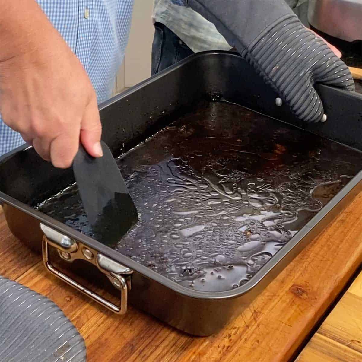 A person using a silicone spatula to scrape up baby back ribs pieces that are stuck to the bottom of a roasting pan that has liquid in it.