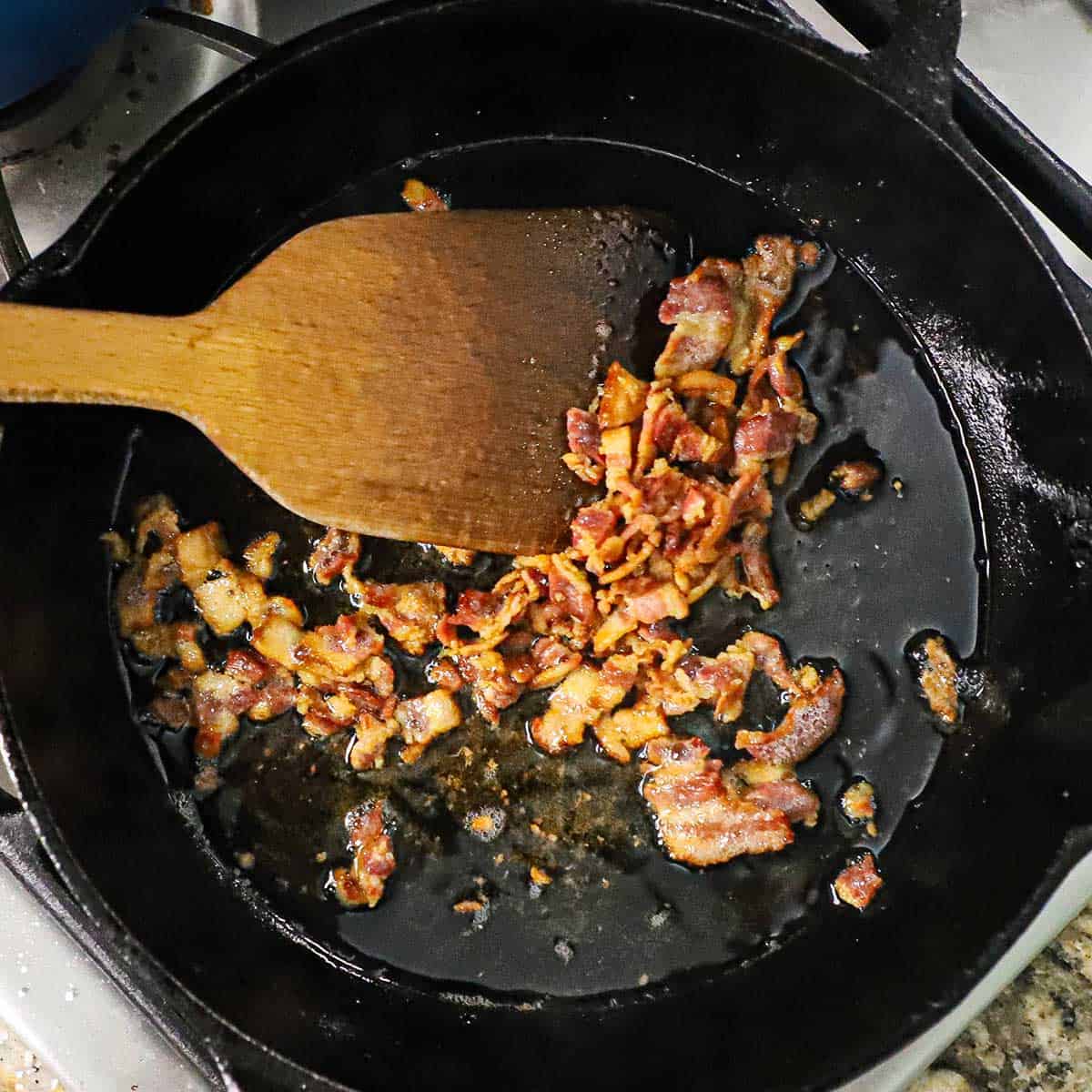 Crumbled bacon being cooked until crisp in a black cast-iron skillet with the bacon being stirred with a large wooden spatula.