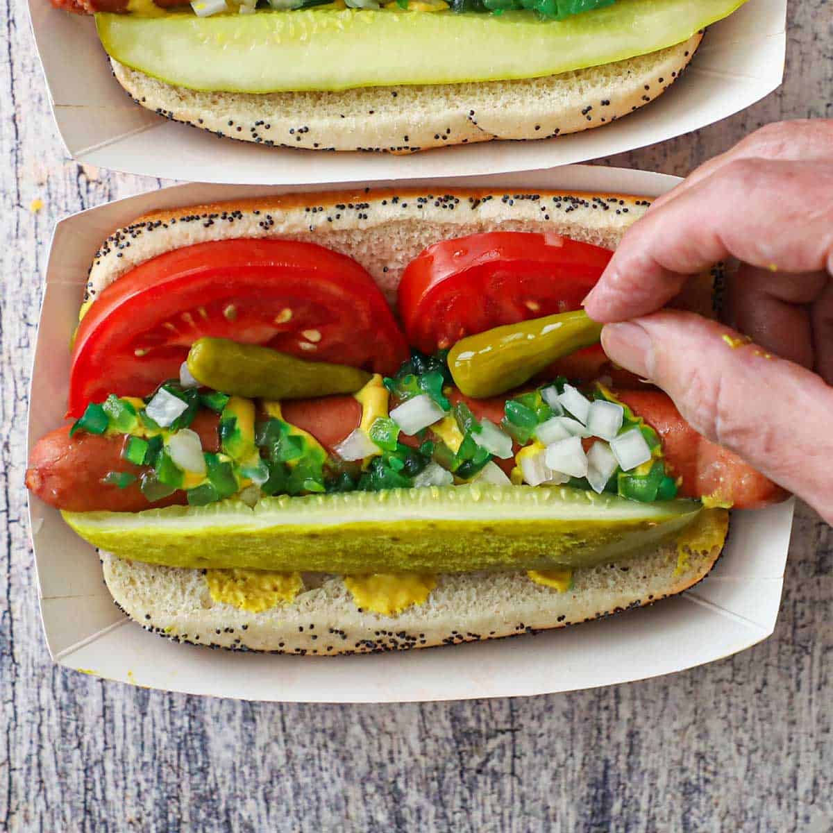 A person placing one of two sport peppers on top of a hot dog that is nestled with tomato wedges, a dill pickle spear, mustard, green relish, and chopped onions.