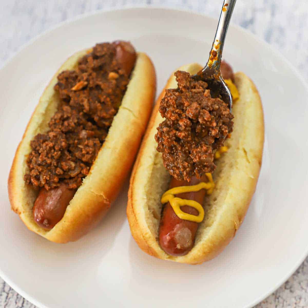 A person using a spoon to add a layer of Cincinnati chili over the tops of two hot dogs that are in hot dog buns with a zig zag layer of mustard on the hot dog.