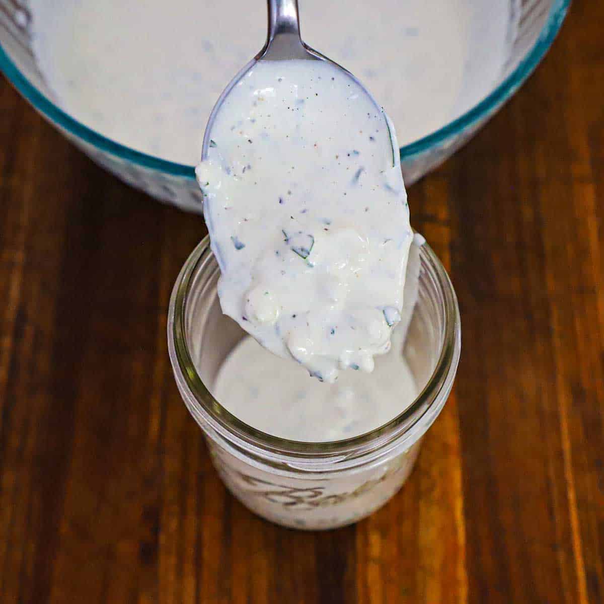 A spoon being used to transfer homemade blue cheese dressing from a large glass bowl into a glass jar on a wooden cutting board.