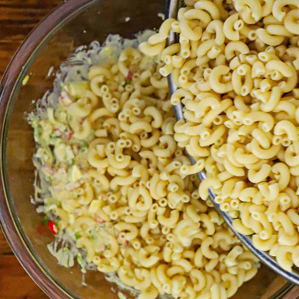 A bowl filled with cooked elbow macaroni being transferred into another large glass bowl filled with a dressing for classic macaroni salad.