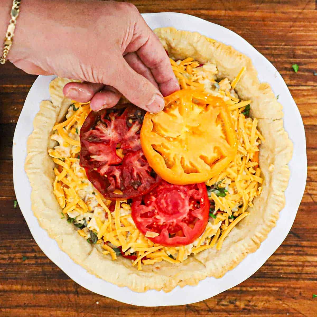 A person placing slices from a red, orange, and yellow heirloom tomato on top of an un-baked Southern tomato pie.