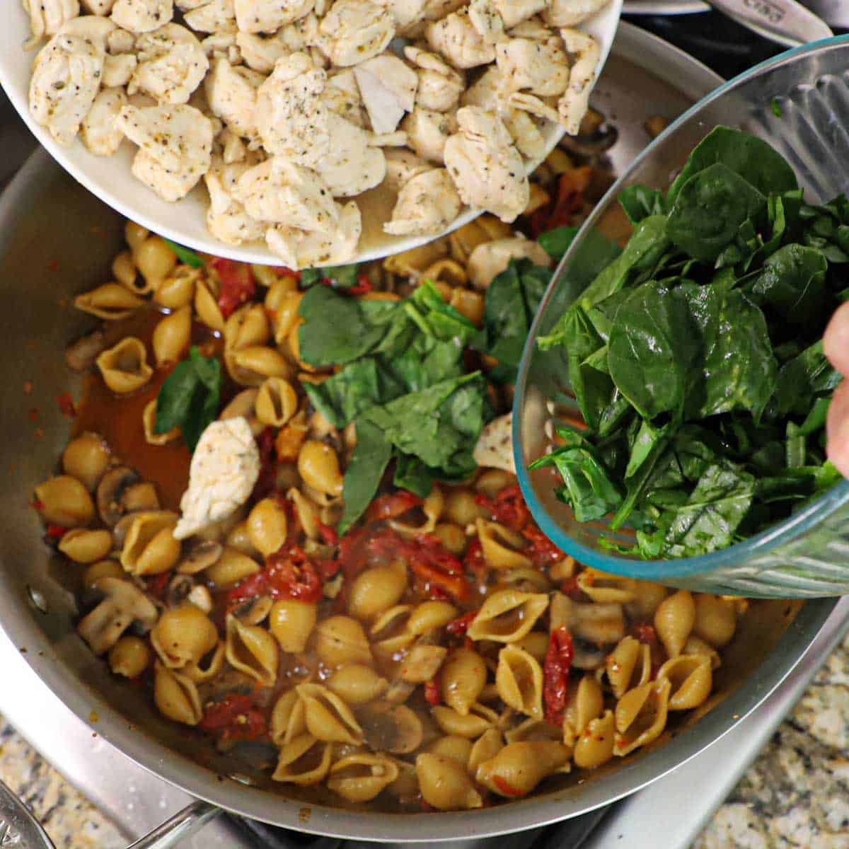 A person holding two glass bowls, one filled with cooked cubed chicken and the other with spinach leaves, with both being transferred into a skillet filled with a simmering Tuscan sauce with pasta and chicken.