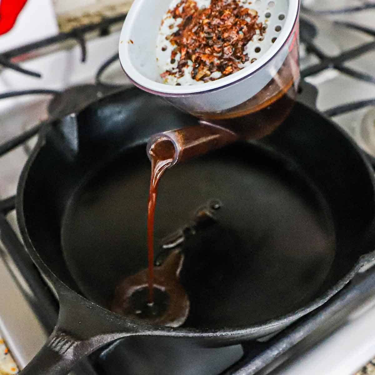 A person pouring a brown liquid from a fat separator into a black cast-iron skillet on the grate of a gas grill.