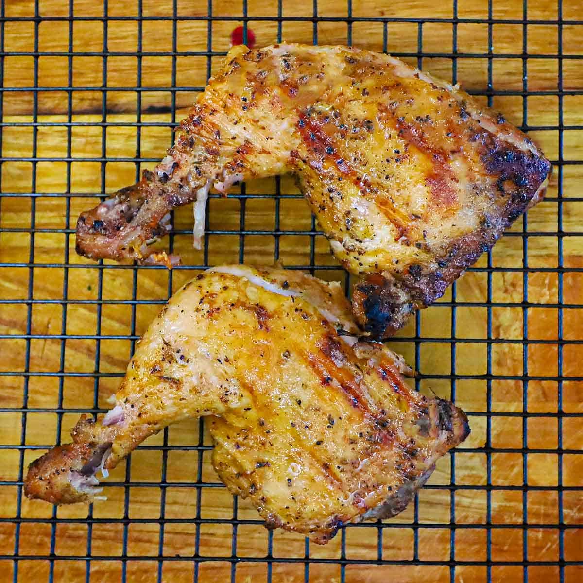 Two fully cooked grilled chicken leg quarters on a baking rack sitting on a wooden cutting board.