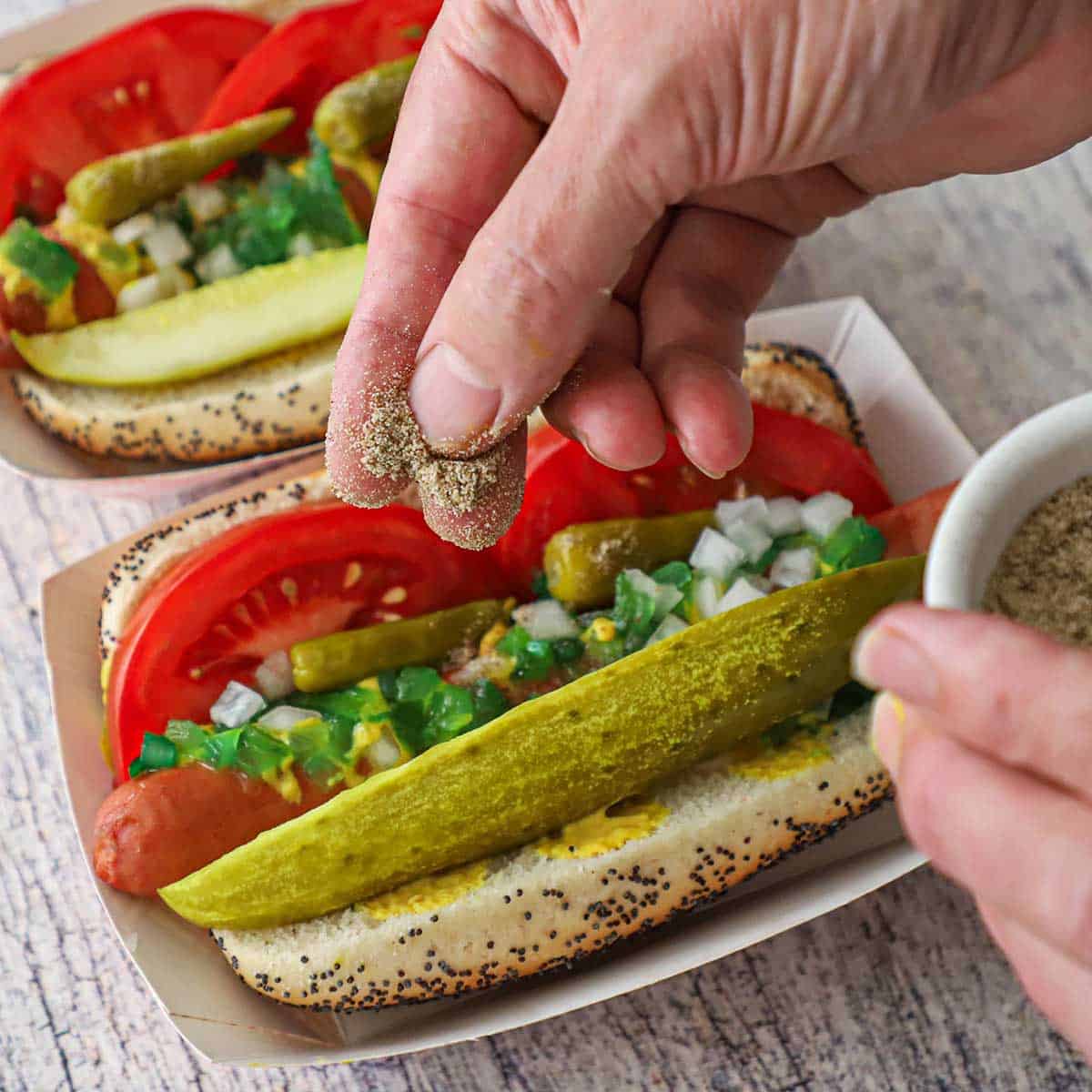 A person using his fingers to sprinkle celery salt over the top of a fully loaded Chicago-style hot dog in a small paper hot dog holder.