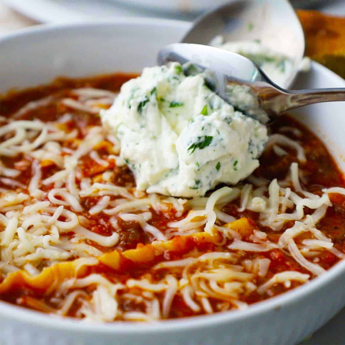 A person using a small spoon to transfer a dollop of ricotta cheese mixed with chopped parsley into a bowl filled with slow cooker lasagna soup.