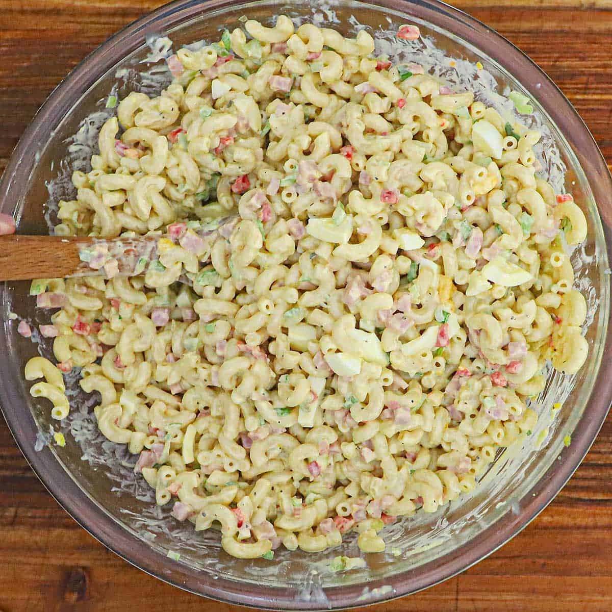 An overhead view of a large glass bowl filled with a classic macaroni salad with a wooden spoon inserted into the salad.