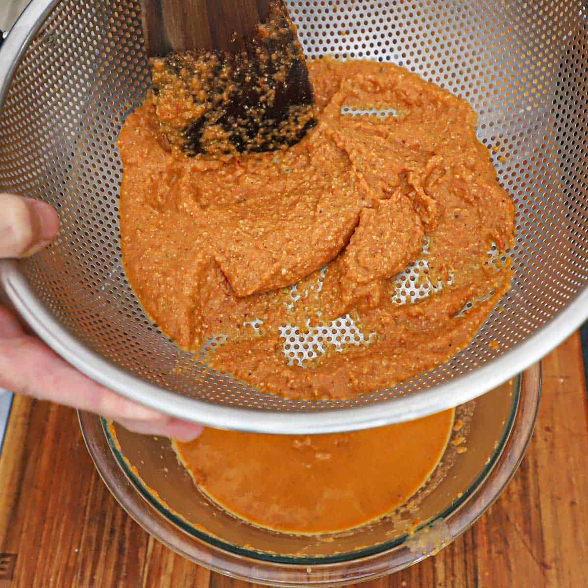 A person using a wooden spatula to pass a puréed butter masala gravy through a colander into a glass bowl underneath it.