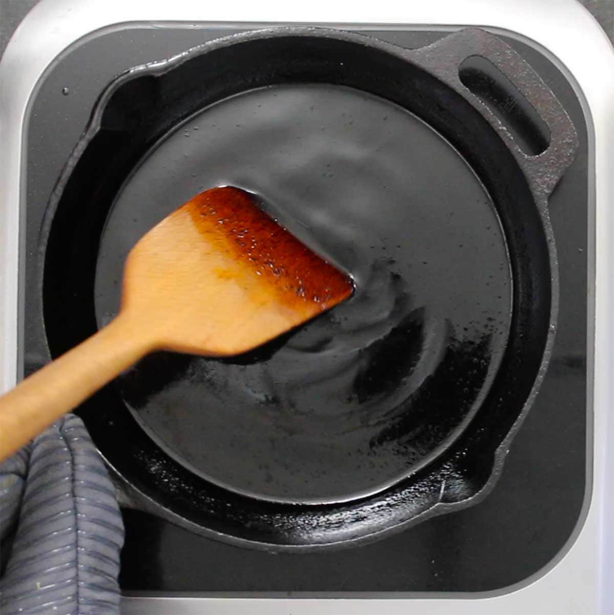 A person using a large wooden spatula to stir a balsamic glaze in a black cast-iron skillet on a hot pad.