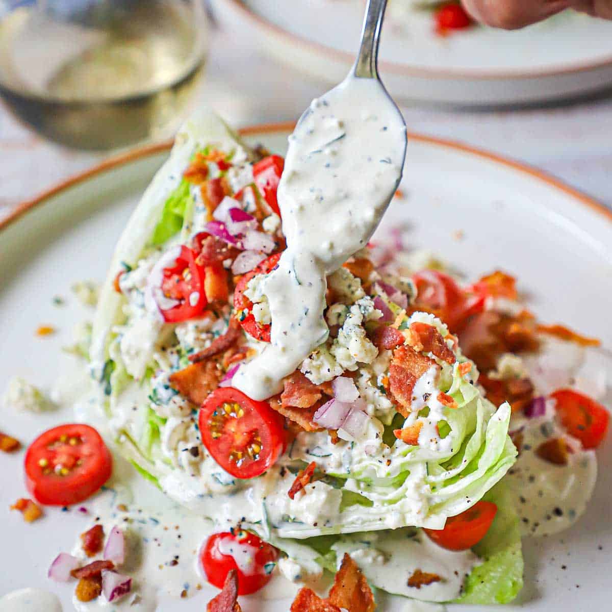 A person using a spoon to drizzle homemade blue cheese dressing over the top of a dressed steakhouse wedge salad.