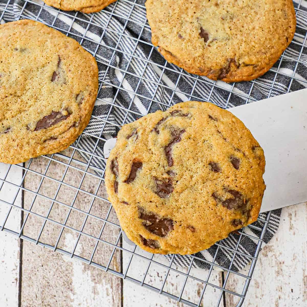 A large metal spatula being used to place a bakery-style chocolate chip cookie onto a baking rack with other cookies already on the rack.