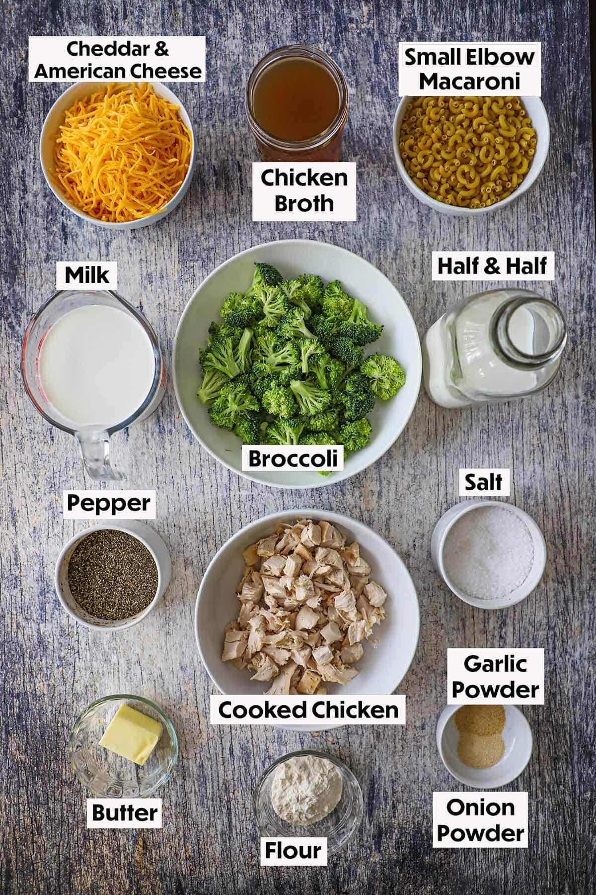 An arrangement of ingredients for creamy mac and cheese with chicken and broccoli on a wooden grey background including shredded cheese, chicken broth, pasta, milk, half and half, broccoli, chicken, butter, and seasonings.