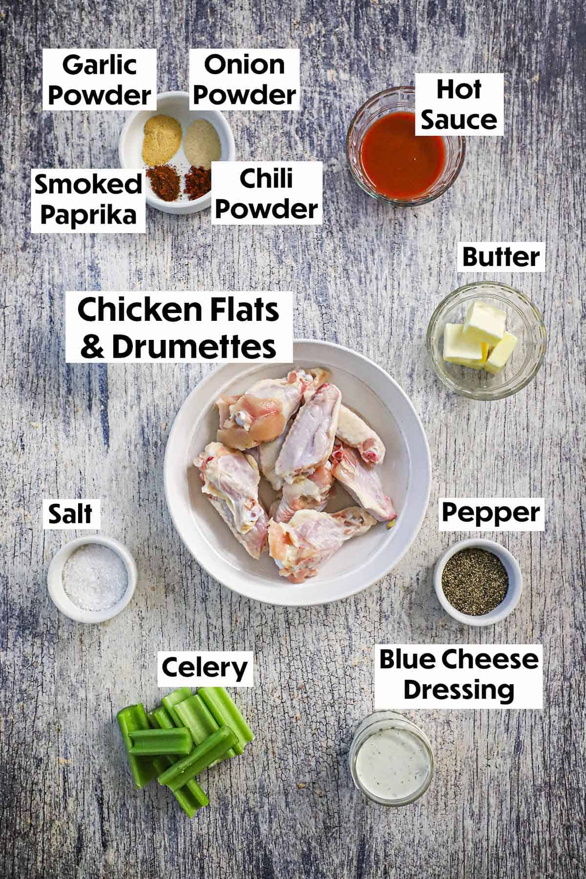 An arrangement of ingredients for grilled chicken wings with buffalo sauce on a wooden background including chicken wings, seasonings, butter, hot sauce, celery, and blue cheese dressing.