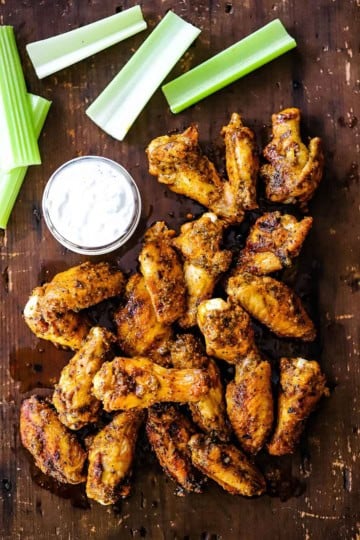 A pile of grilled chicken wings with buffalo sauce resting on a wooden board next to a jar of blue cheese dressing and cut strips of celery.