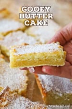A person holding a square piece of a Gooey Butter Cake that has been dusted with powdered sugar and is near other cut squares of the cake.