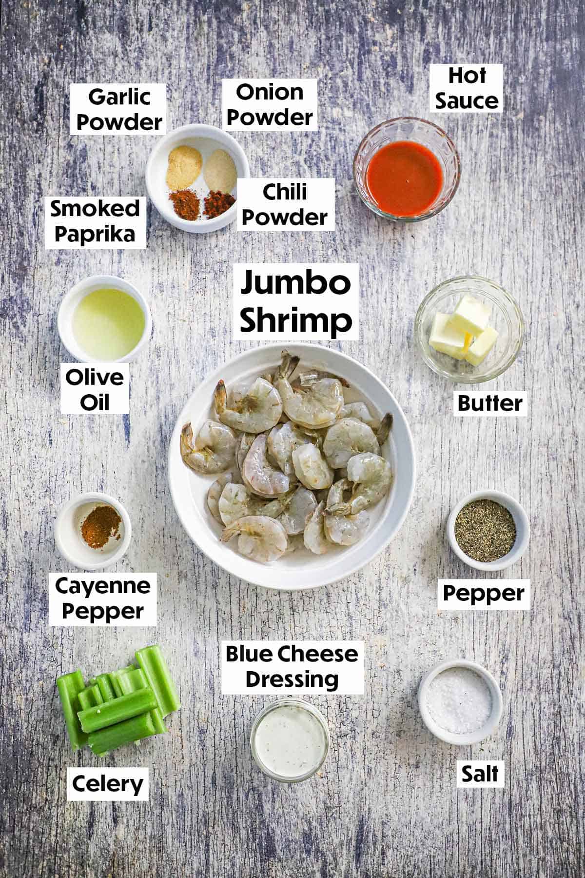 An arrangement of ingredient for Grilled Buffalo Shrimp on a wooden grey background including uncooked shrimp, seasonings, hot sauce, butter, olive oil, blue cheese dressing, and celery.