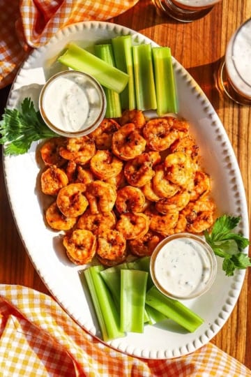 A white oval platter filled with a pile of grilled buffalo shrimp with two small jars filled with blue cheese dressing and celery sticks on each side of the dish.