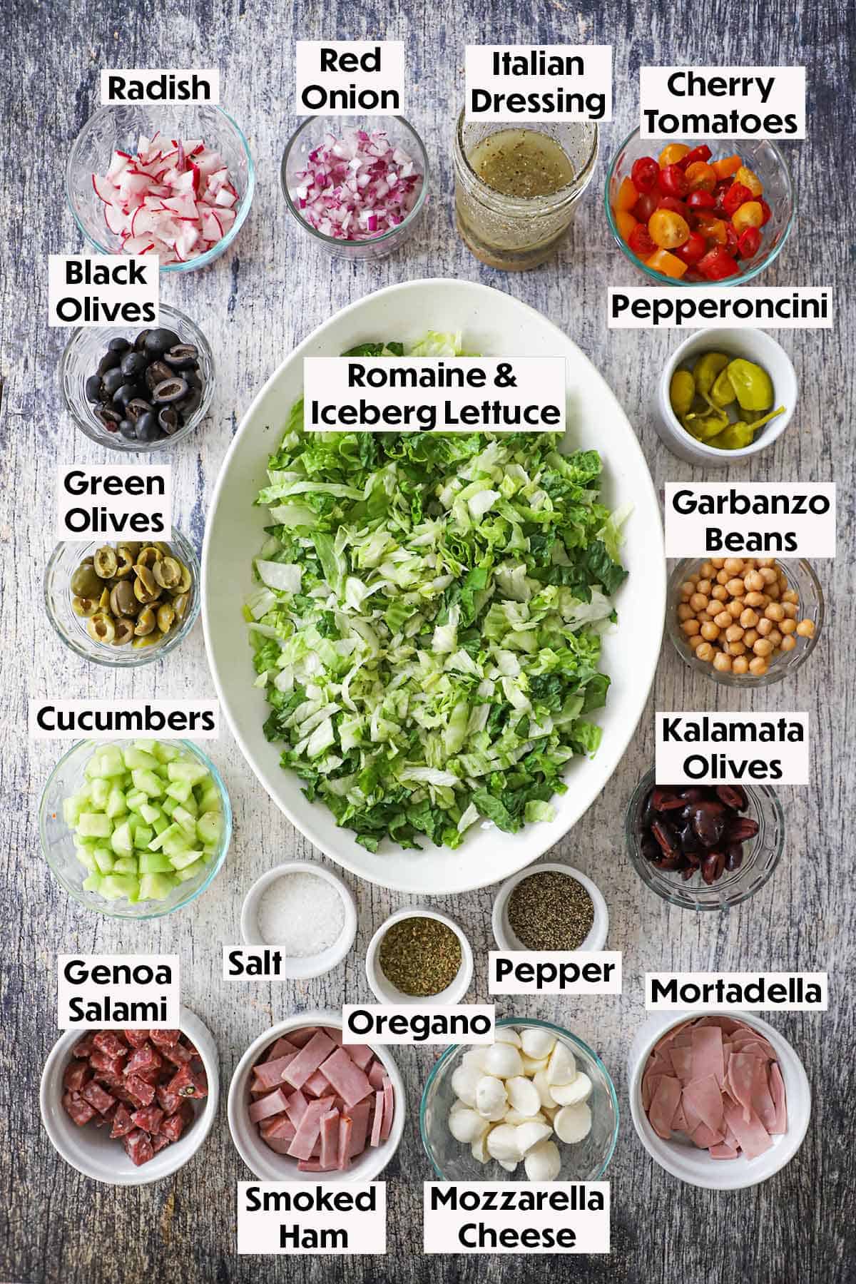 An arrangement of ingredients on a grey wooden background for Italian chopped salad with homemade vinaigrette including chopped lettuce, vegetables, Italian cured meats, Mozzarella cubes, olives, Italian dressing, and seasonings.