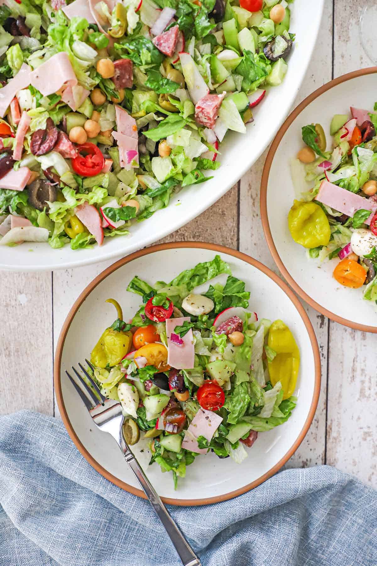 Two salad plates filled with individual servings of Italian chopped salad with homemade vinaigrette next to a large platter filled with the same salad.