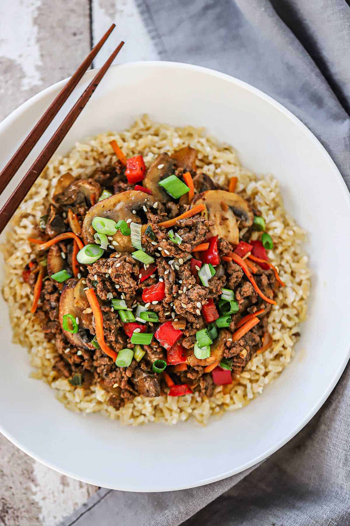 An overhead view of Korean ground beef stir-fry in a white bowl over a bed of steamed brown rice with a pair of brown chop sticks resting on the side of the bowl.