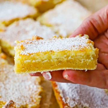 A person holding a square piece of a Gooey Butter Cake that has been dusted with powdered sugar and is near other cut squares of the cake.