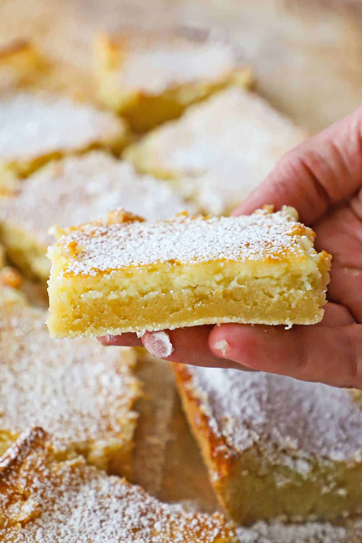 A person holding a square piece of a Gooey Butter Cake that has been dusted with powdered sugar and is near other cut squares of the cake.