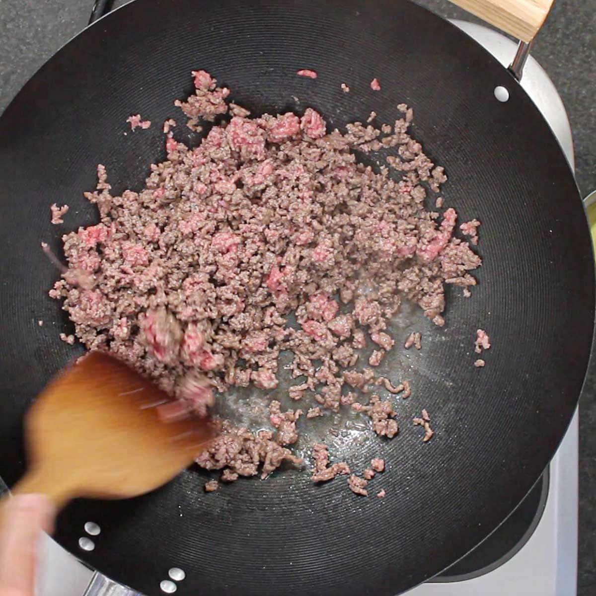A person using a wooden spatula to stir simmering ground beef that is being cooked in a wok.
