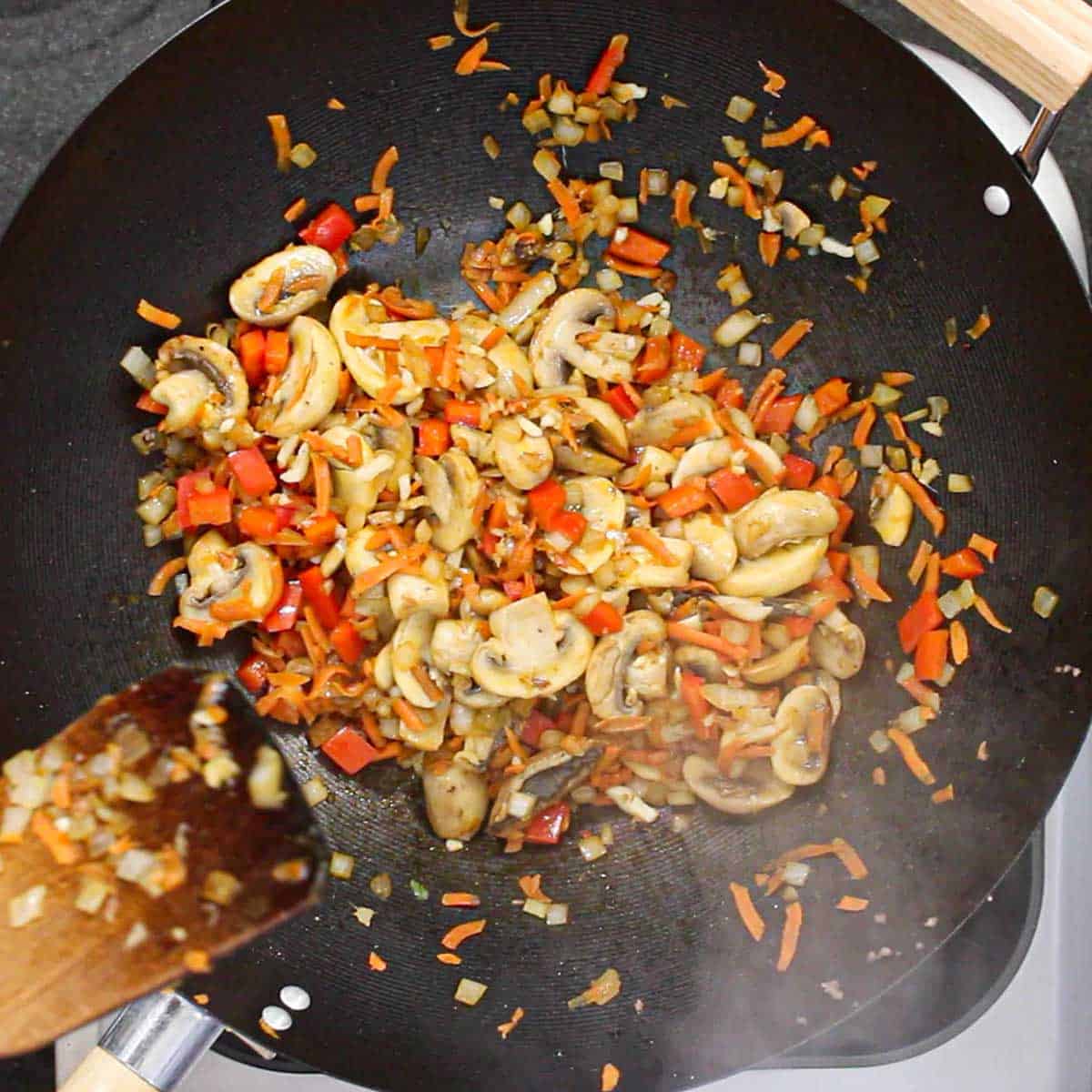 A person using a wooden spatula to sauté sliced mushrooms, chopped bell peppers, and shredded carrots in a large black wok.