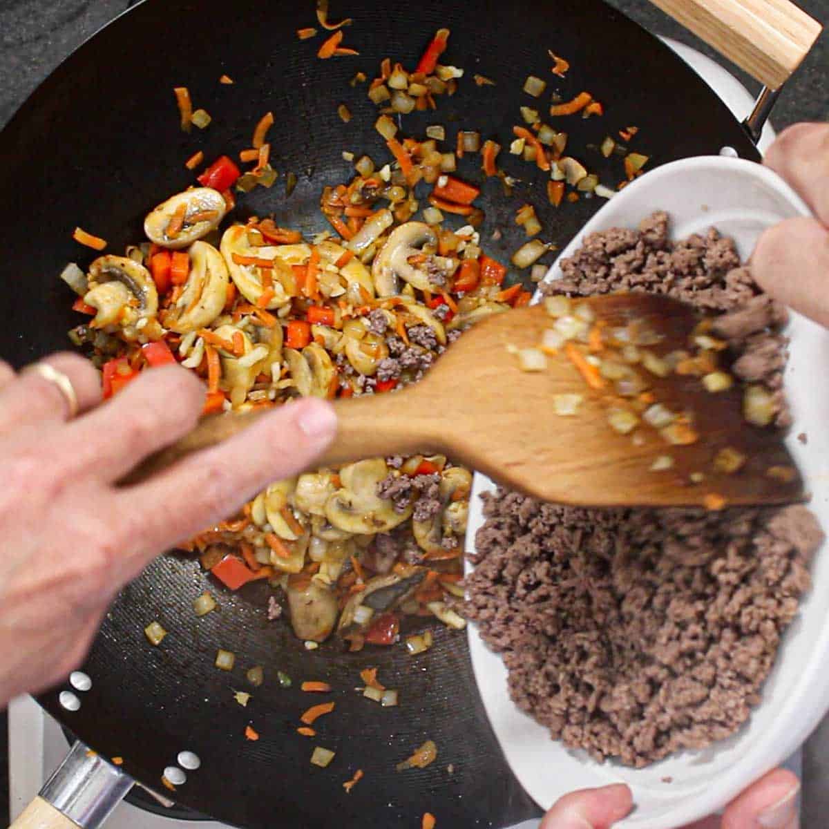 A person using a wooden spatula to transfer cooked ground beef from a white bowl into a wok filled with simmering sautéed mushrooms, bell peppers, and shredded carrots.