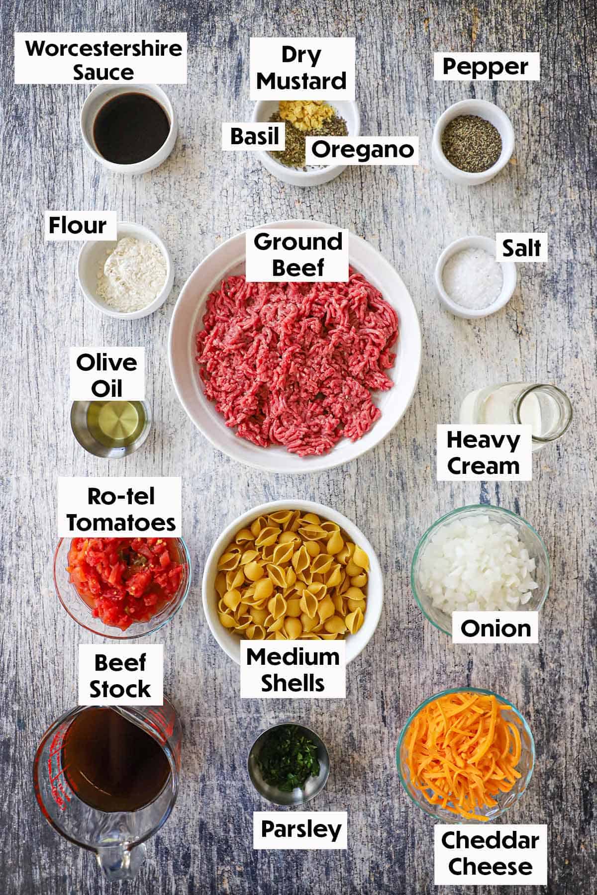 An arrangement of ingredients for creamy beef and shells on a grey wooden background including ground beef, seasonings, flour, chopped tomatoes, pasta shells, onion, cheddar cheese, onion, and beef stock.
