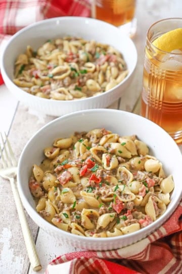 Two white pasta bowls filled with servings of creamy beef and shells with a glass of ice tea and lemon nearby.