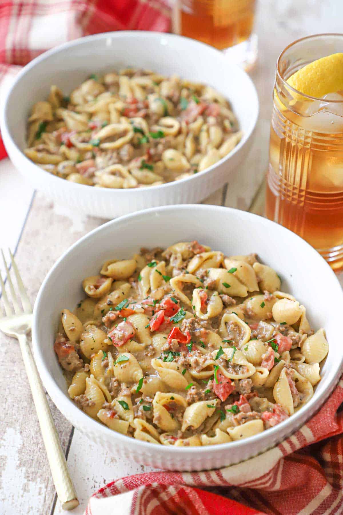 Two white pasta bowls filled with servings of creamy beef and shells with a glass of ice tea and lemon nearby.