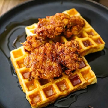 A serving of crispy chicken and waffles resting on a black plated with a fork and a glass mug of coffee nearby.