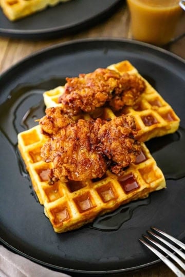 A serving of crispy chicken and waffles resting on a black plated with a fork and a glass mug of coffee nearby.