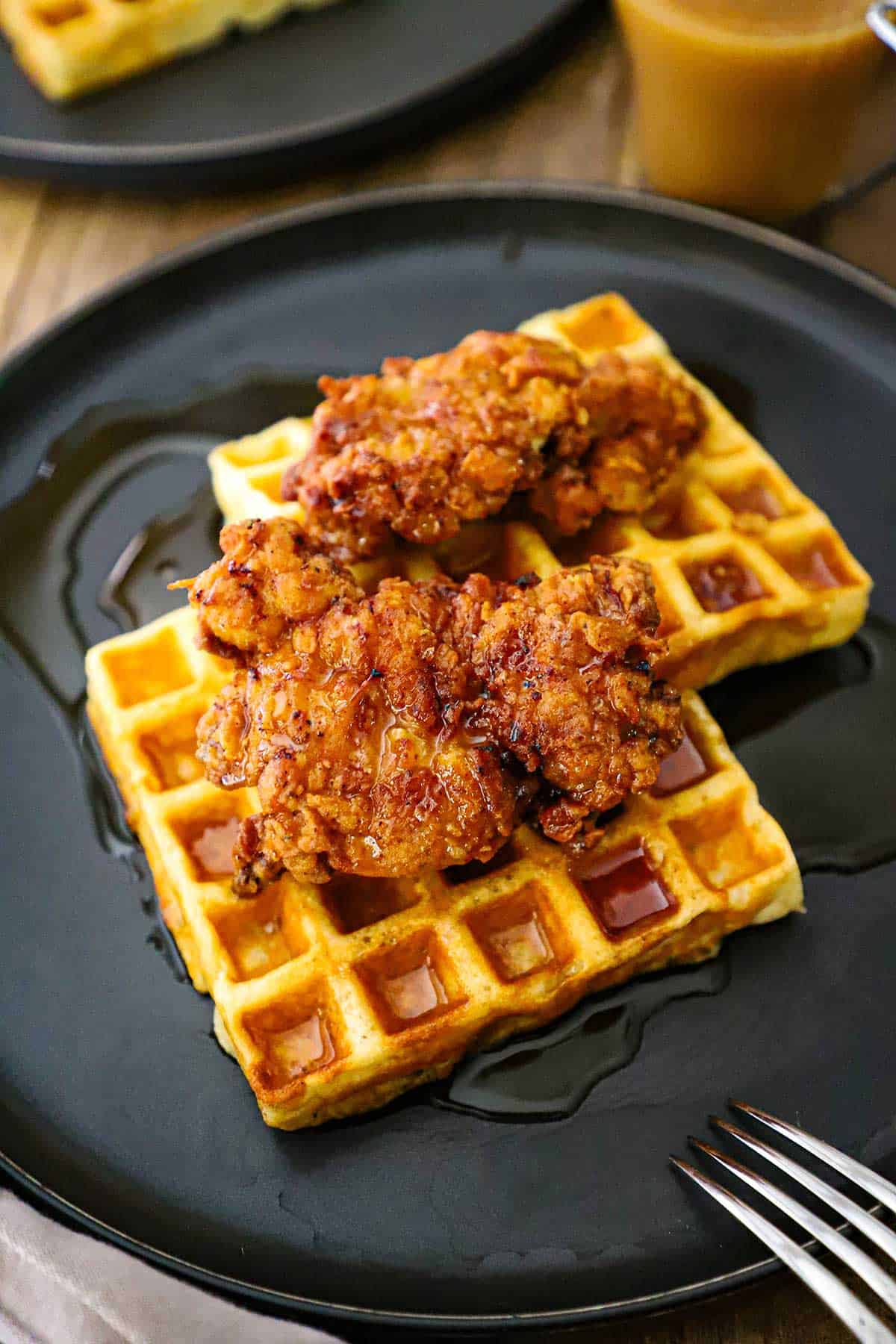 A serving of crispy chicken and waffles resting on a black plated with a fork and a glass mug of coffee nearby.