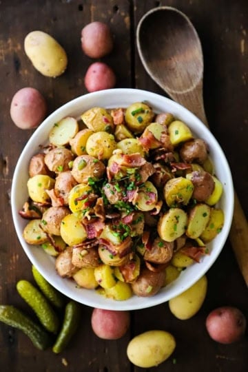 An overhead view of a white serving bowl filled with a mound of Best German Potato Salad that is topped with chopped crispy bacon and snipped chives.