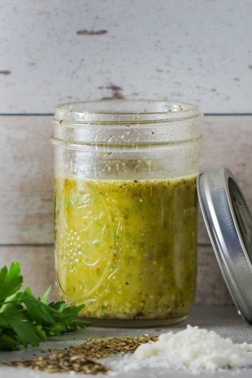 A Mason jar filled with homemade Italian dressing with a small pile of grated parmesan cheese, dried herbs, and fresh parsley around the jar.