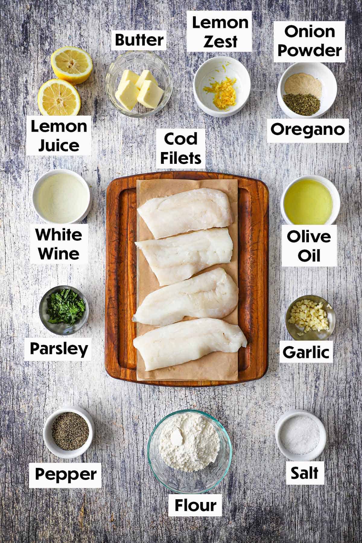 An arrangement of ingredient for pan-fried cod with lemon butter sauce on a wooden grey background including cod filets, butter, lemon, white wine, garlic, flour, Italian parsley, and seasonings.