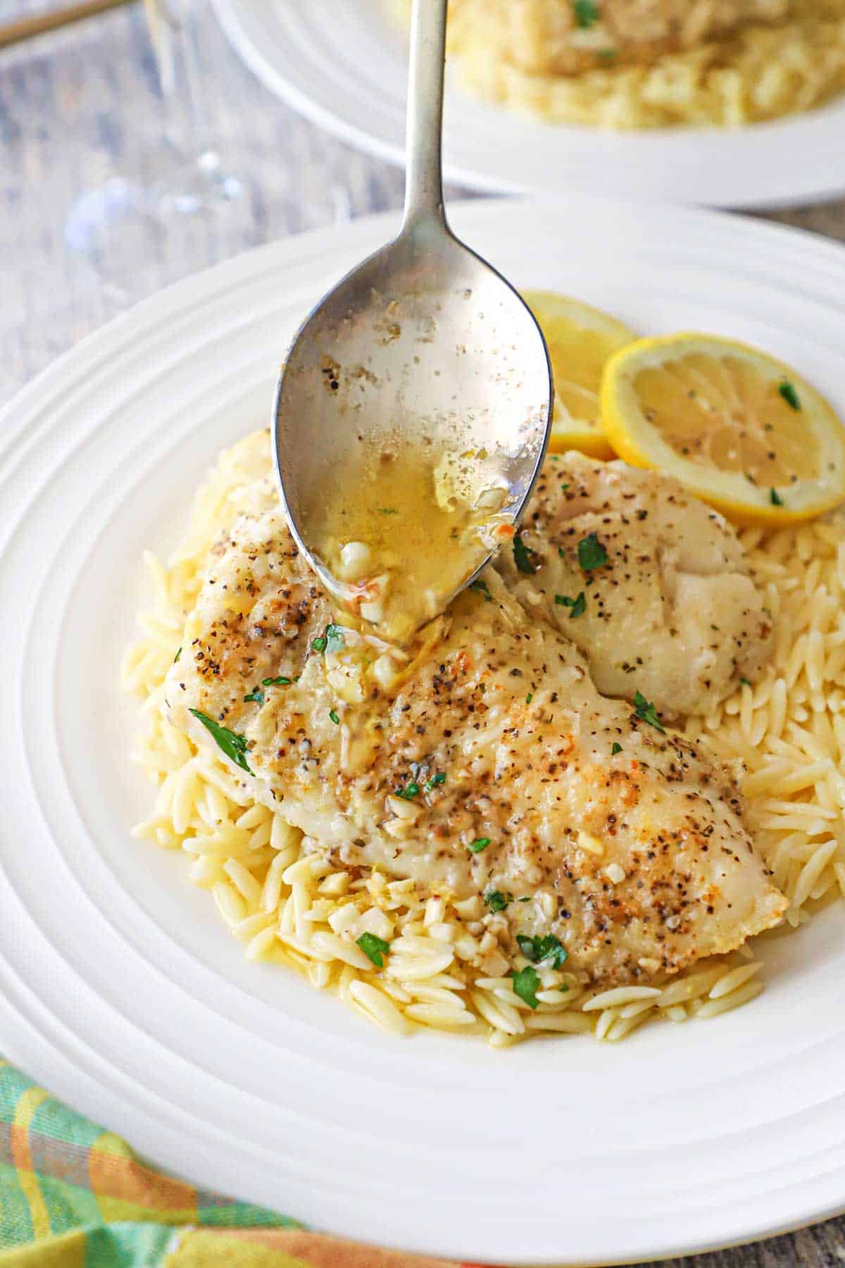 A person using a serving spoon to drizzle a lemon butter sauce over two pan-fried filets of cod on a bed of orzo on a white dinner plate.