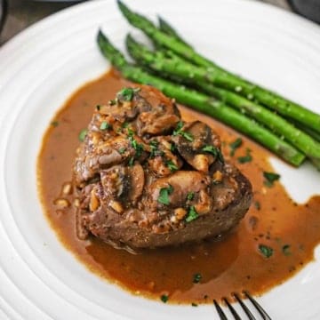 A serving of Classic Steak Diane on a white dinner plate with sautéed mushrooms on top of the filet as well as a brown sauce surrounding the steak and next to roasted asparagus.