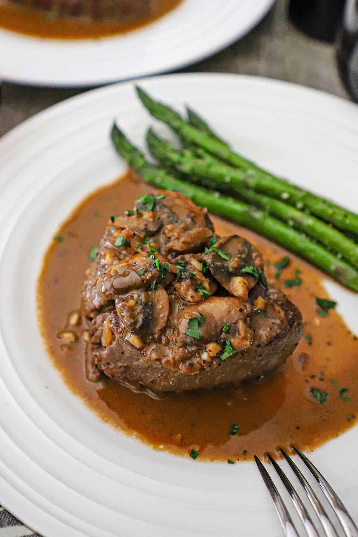 A serving of Classic Steak Diane on a white dinner plate with sautéed mushrooms on top of the filet as well as a brown sauce surrounding the steak and next to roasted asparagus.
