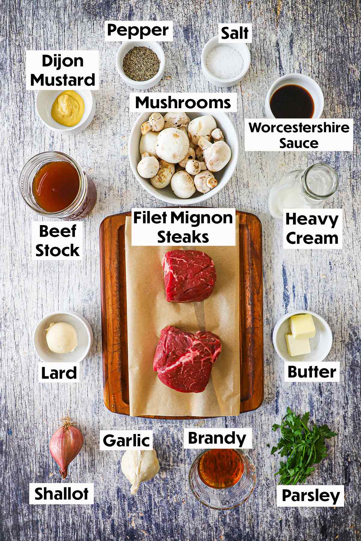 An arrangement of ingredients for Classic Steak Diane on a grey wooden background including filet mignon steaks, mushrooms, Dijon mustard, cream, beef stock, butter, brandy, garlic, a shallot, and parsley.
