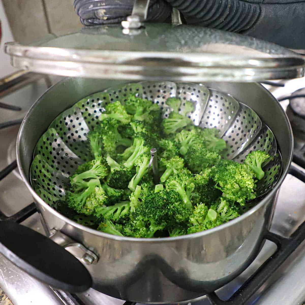 A person lifting a clear lid from a metal pot on a stove that contains a steamer basket with freshly steamed broccoli florets resting on the basket.