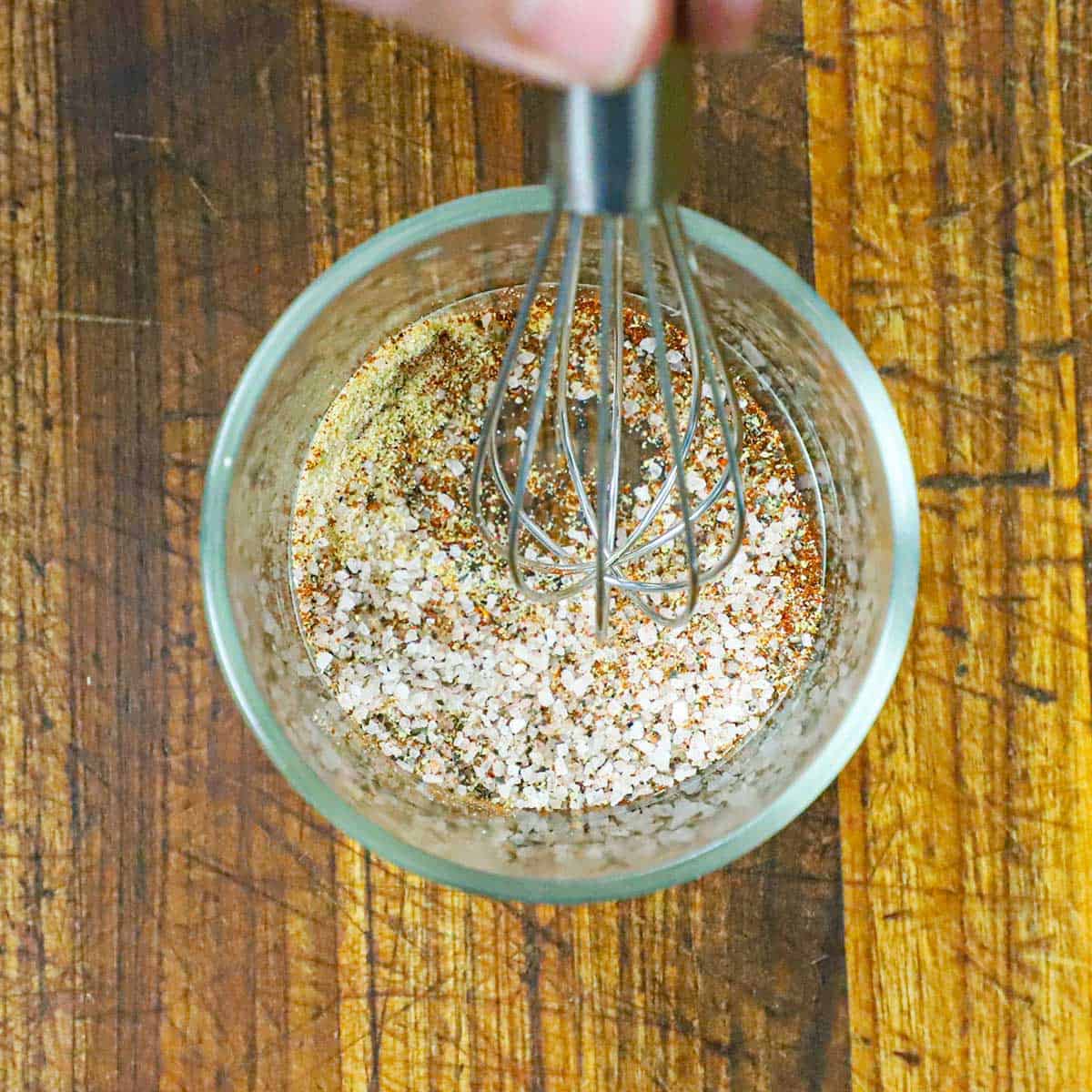 A person using a small whisk to combine salt, chili powder, paprika, and garlic powder in a small glass bowl on a cutting board.