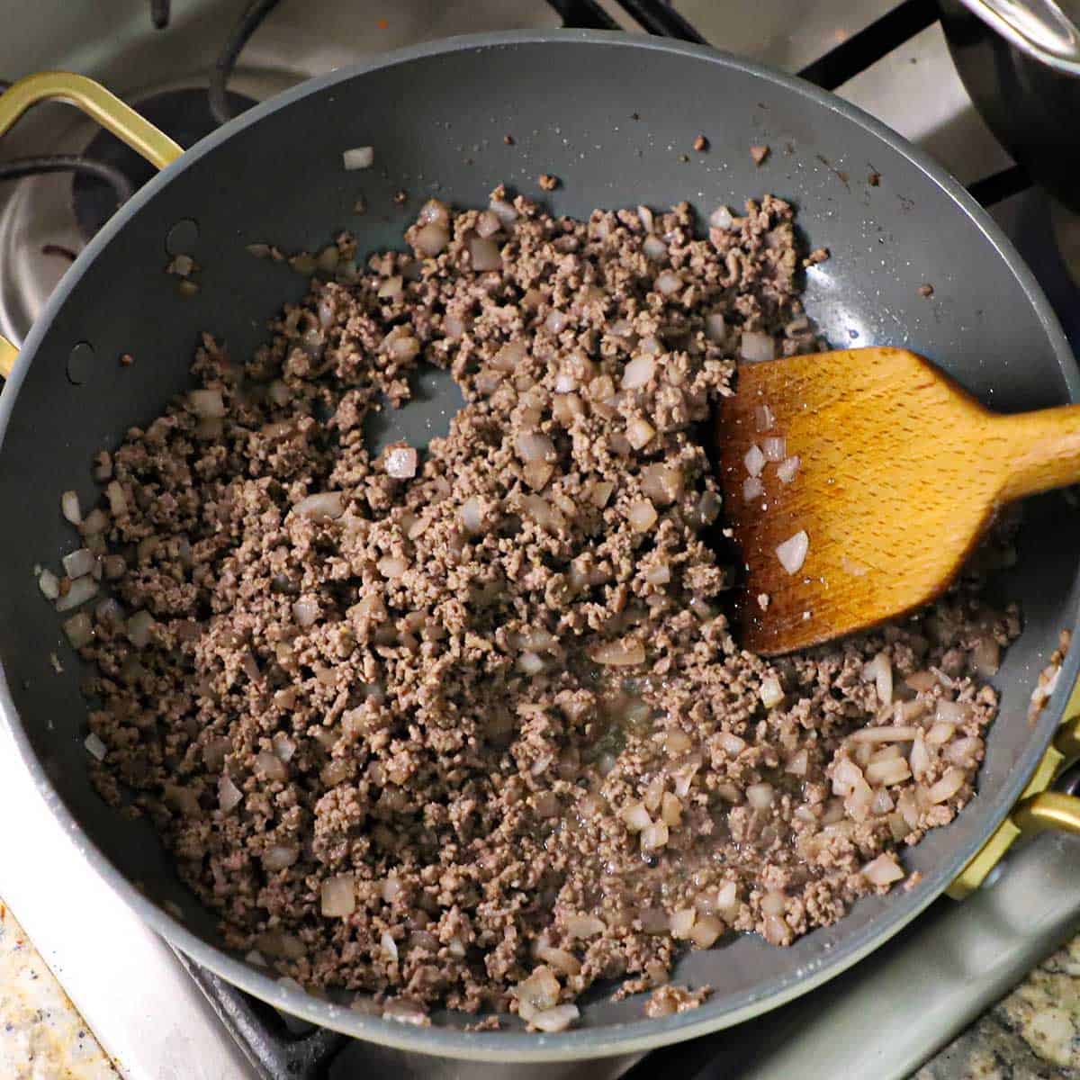 A large skillet filled with cooked ground beef and sautéed onions with a wooden spatula in the meat mixture.