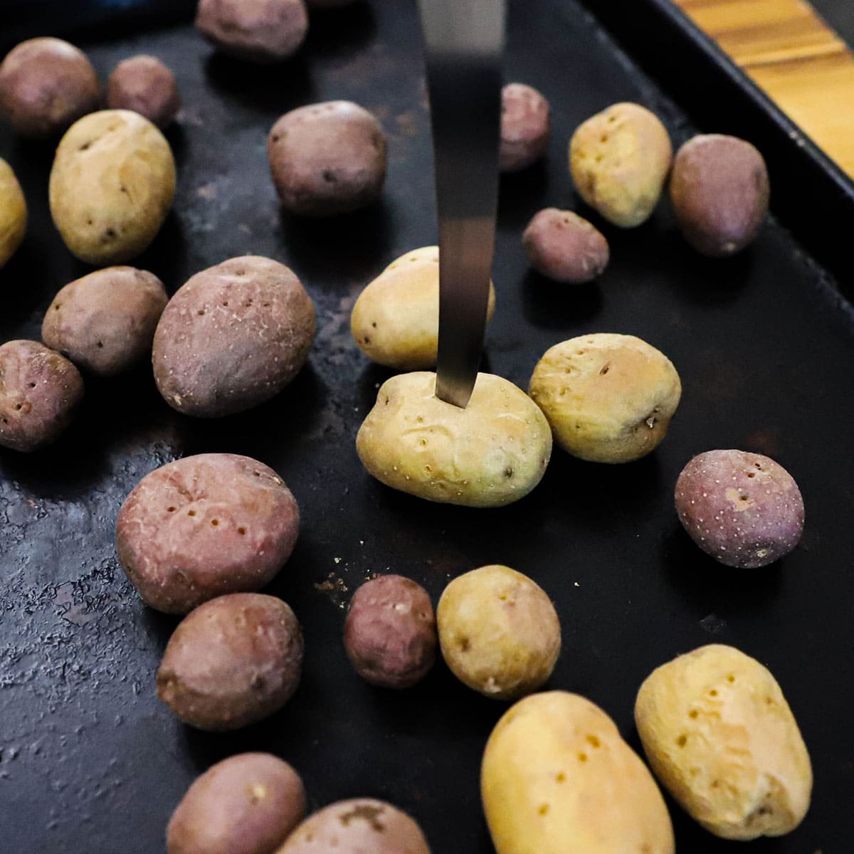 A person using a sharp knife to pierce a cooked baby potato on a baking sheet surround by other cooked potatoes.