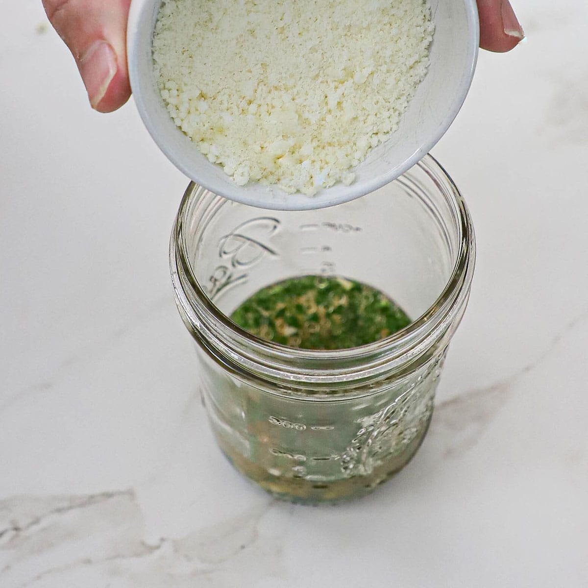 A person dumping grated Parmesan cheese from a small white bowl into a jar filled with white wine vinegar, Italian parsley, lemon juice, and garlic.