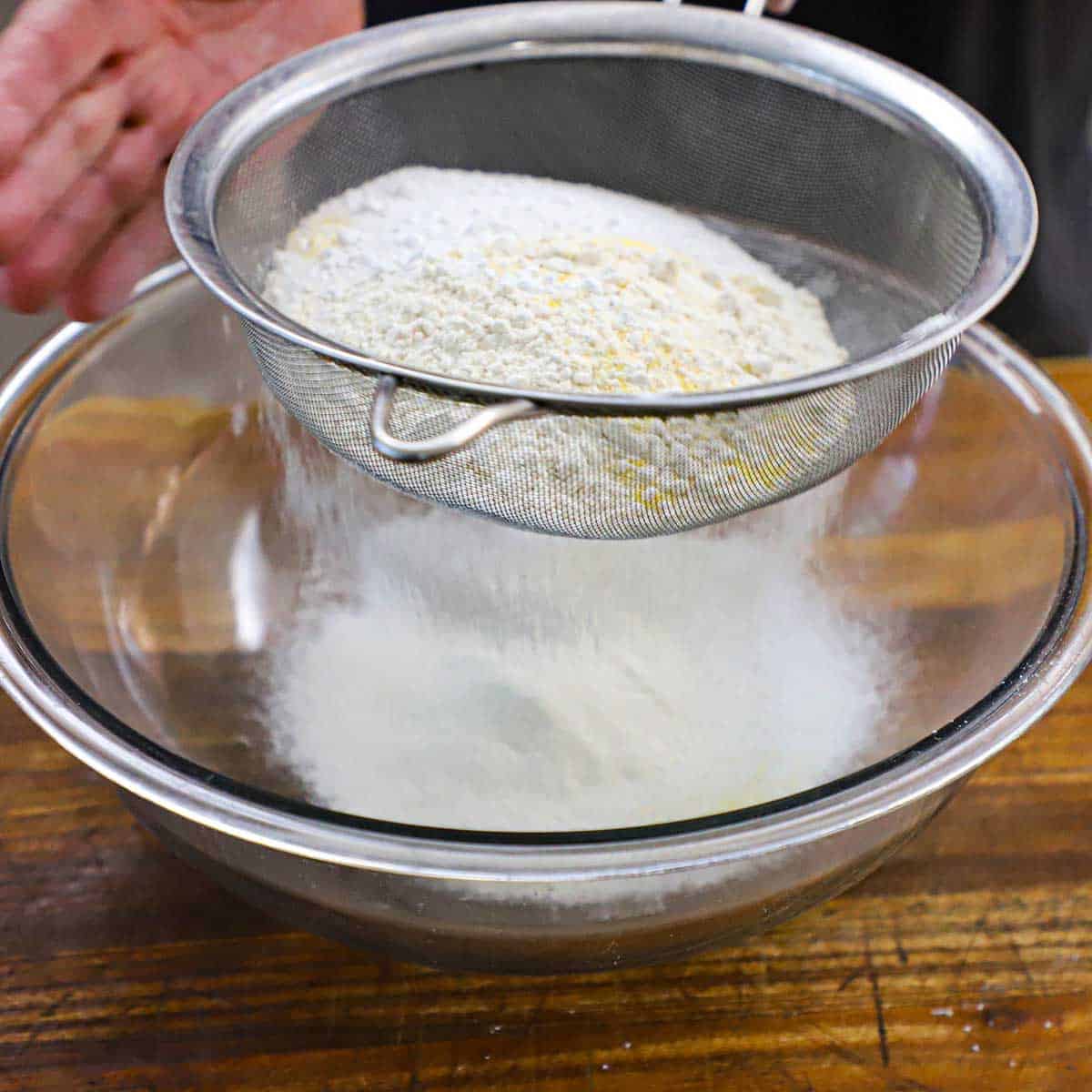 A person using a large sieve to sift flour, sugar, and cornmeal into a large glass bowl resting on a wooden cutting board.